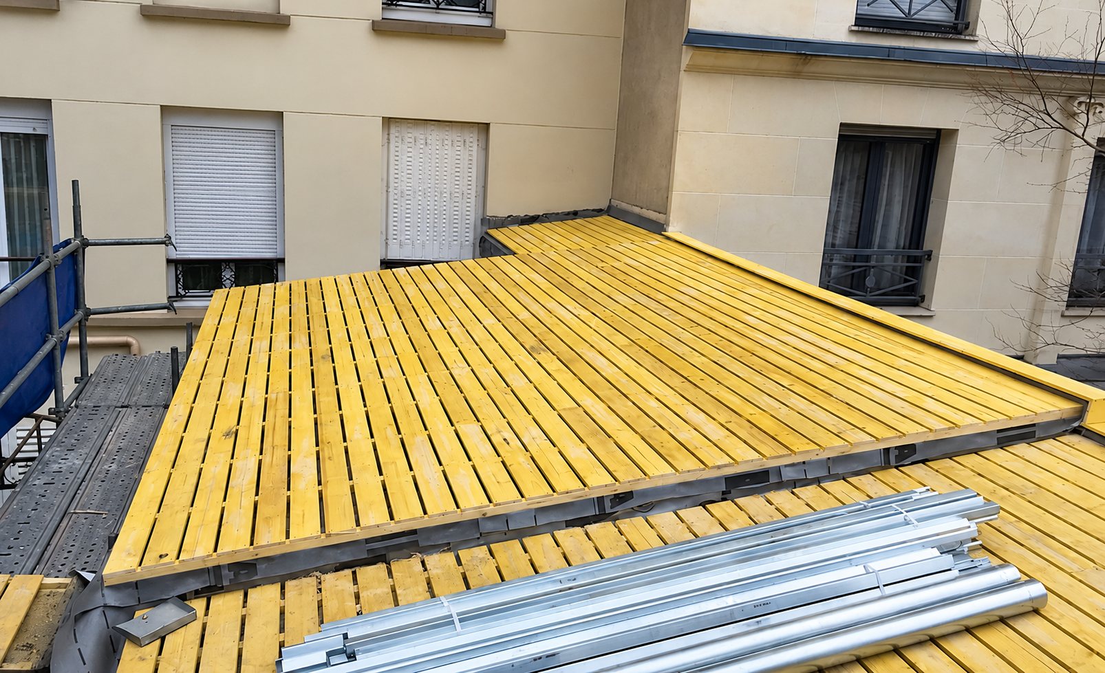 Un toit-terrasse en cours de travaux, recouvert d'un platelage en bois jaune, avec des profilés métalliques posés au premier plan et un échafaudage à gauche entre des façades d'immeubles.