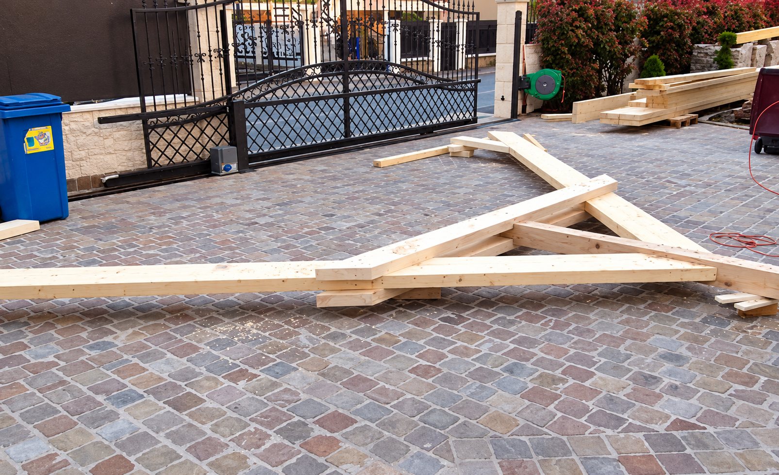 Une ferme de charpente en bois clair est posée à plat sur une allée pavée devant un portail en fer forgé noir et une poubelle bleue.
