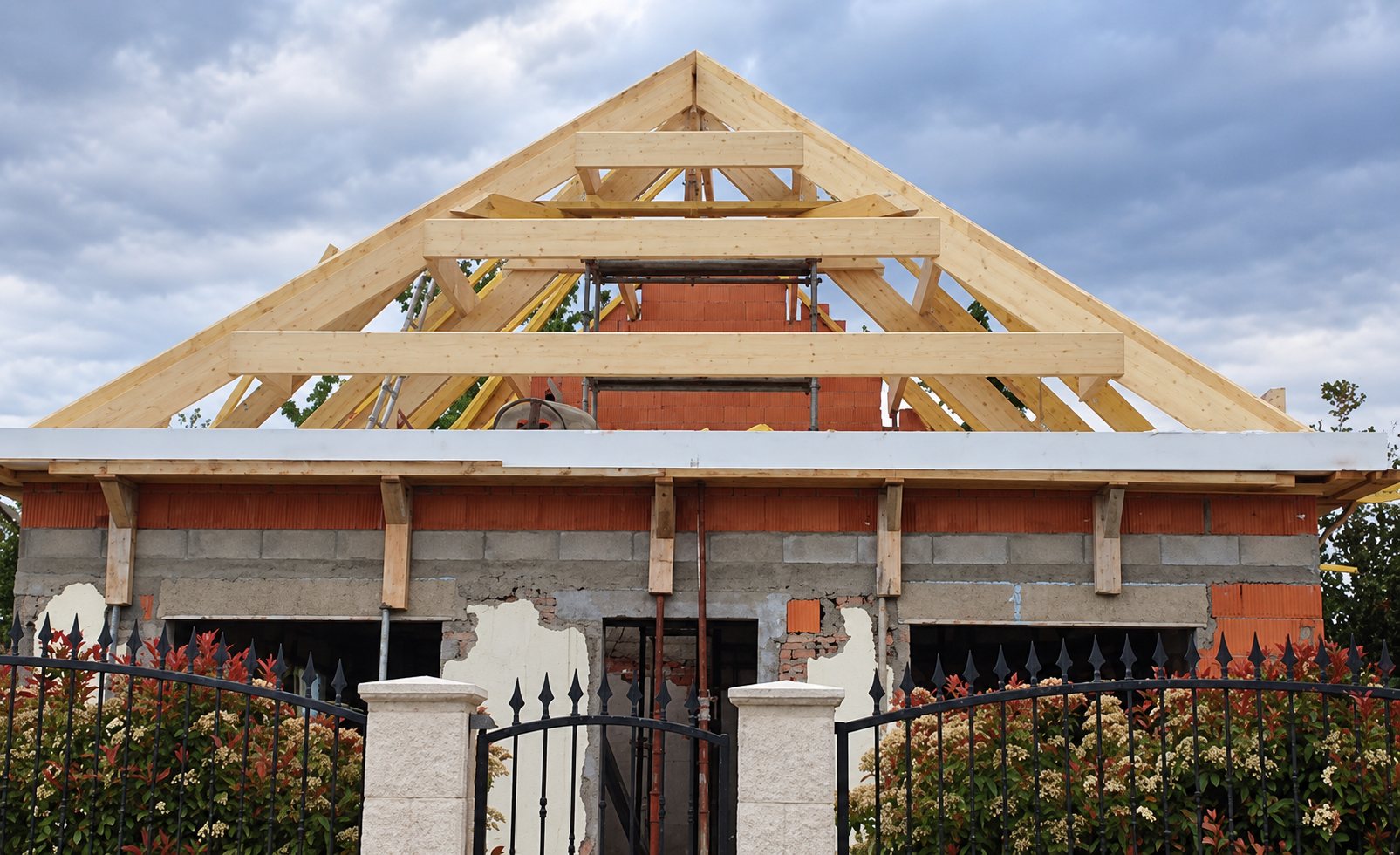 Charpente traditionnelle en bois neuve posée sur les murs en briques d'une maison en construction, derrière un portail et une clôture en fer forgé.