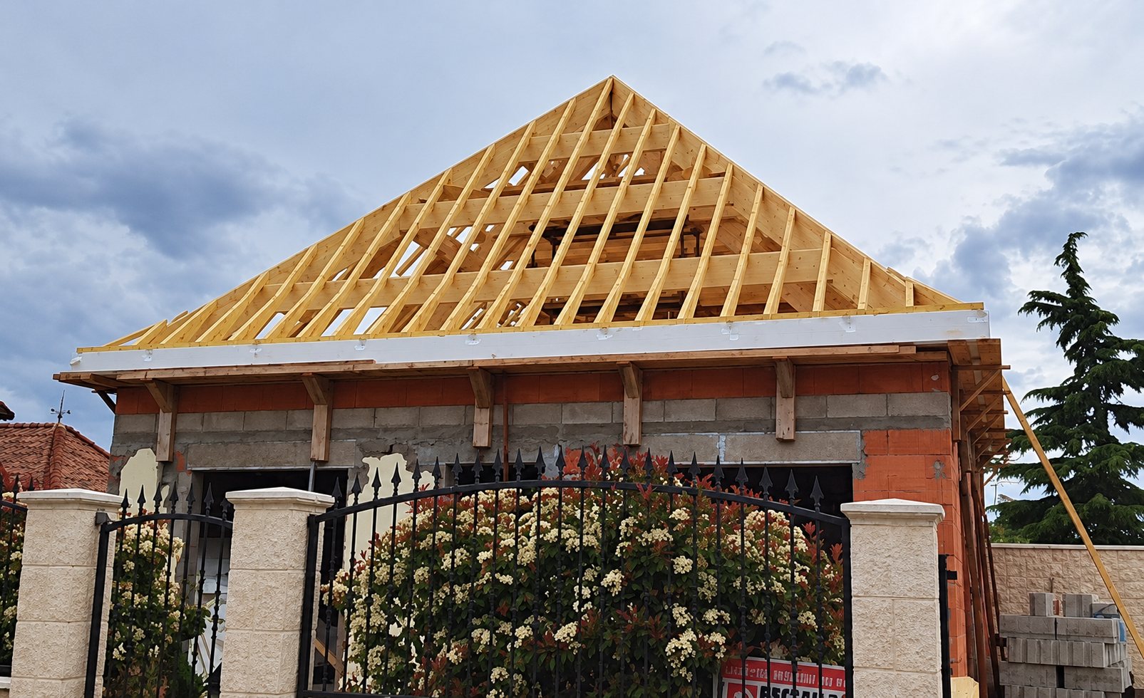 Une charpente en bois clair à quatre pans est posée sur les murs en briques et parpaings d'une maison en chantier, derrière un portail en fer forgé et des rosiers en fleurs.