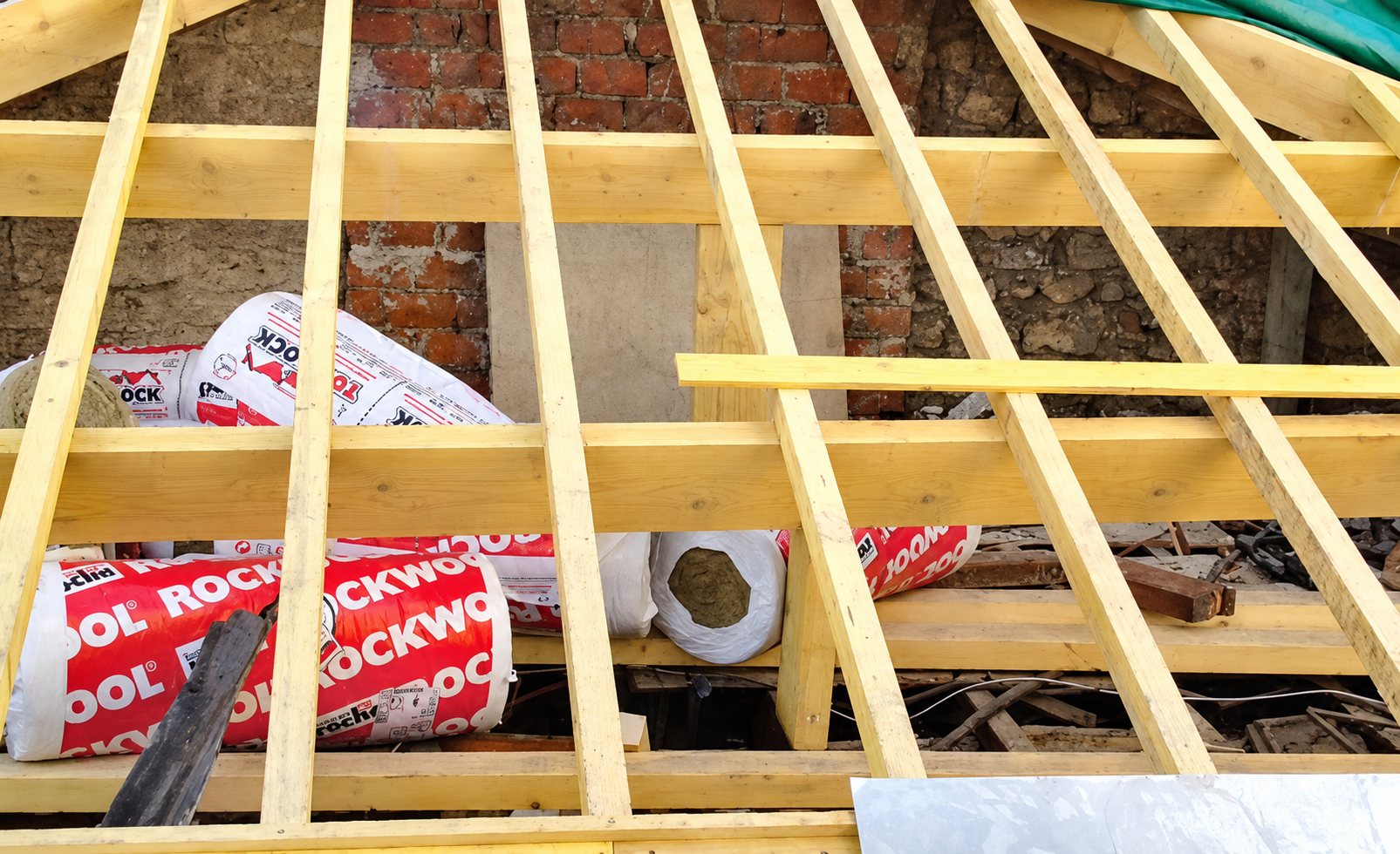 Chevrons de bois neufs d'une charpente en cours de pose, avec des rouleaux d'isolant Rockwool posés entre les poutres devant un mur de briques.