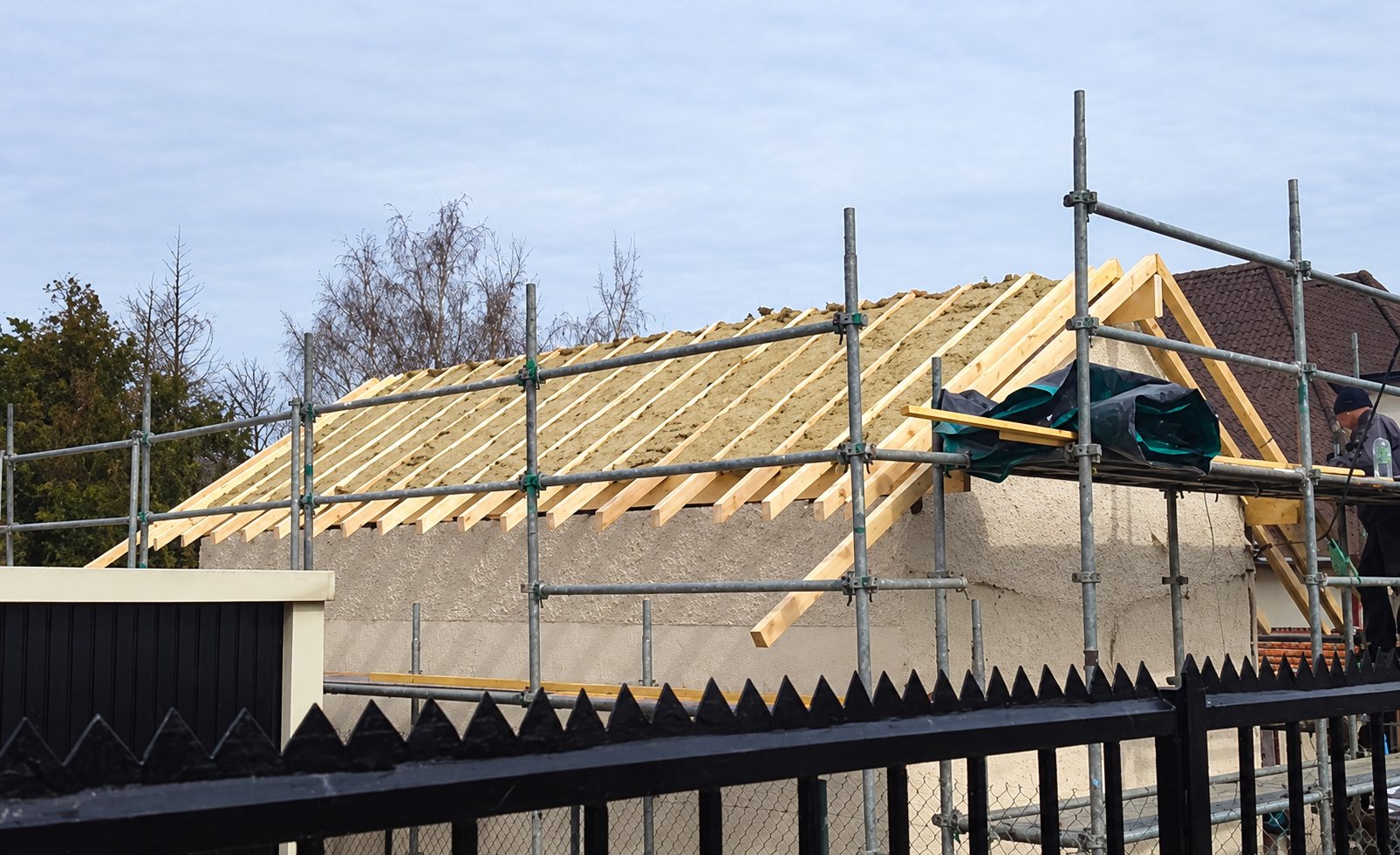 Une charpente en bois en cours de pose sur un bâtiment aux murs clairs, entourée d'un échafaudage métallique et d'une clôture noire au premier plan.