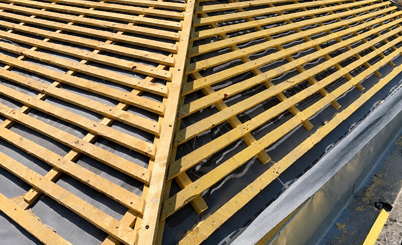 Vue rapprochée d'une toiture en construction avec des liteaux en bois posés sur un écran de sous-toiture noir, de part et d'autre d'une arête.