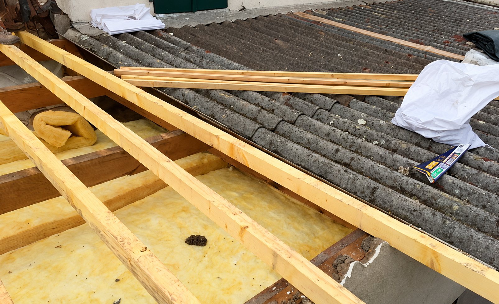 Des chevrons en bois neufs sont posés sur une toiture en plaques ondulées grises, avec de la laine de verre jaune visible entre les poutres.