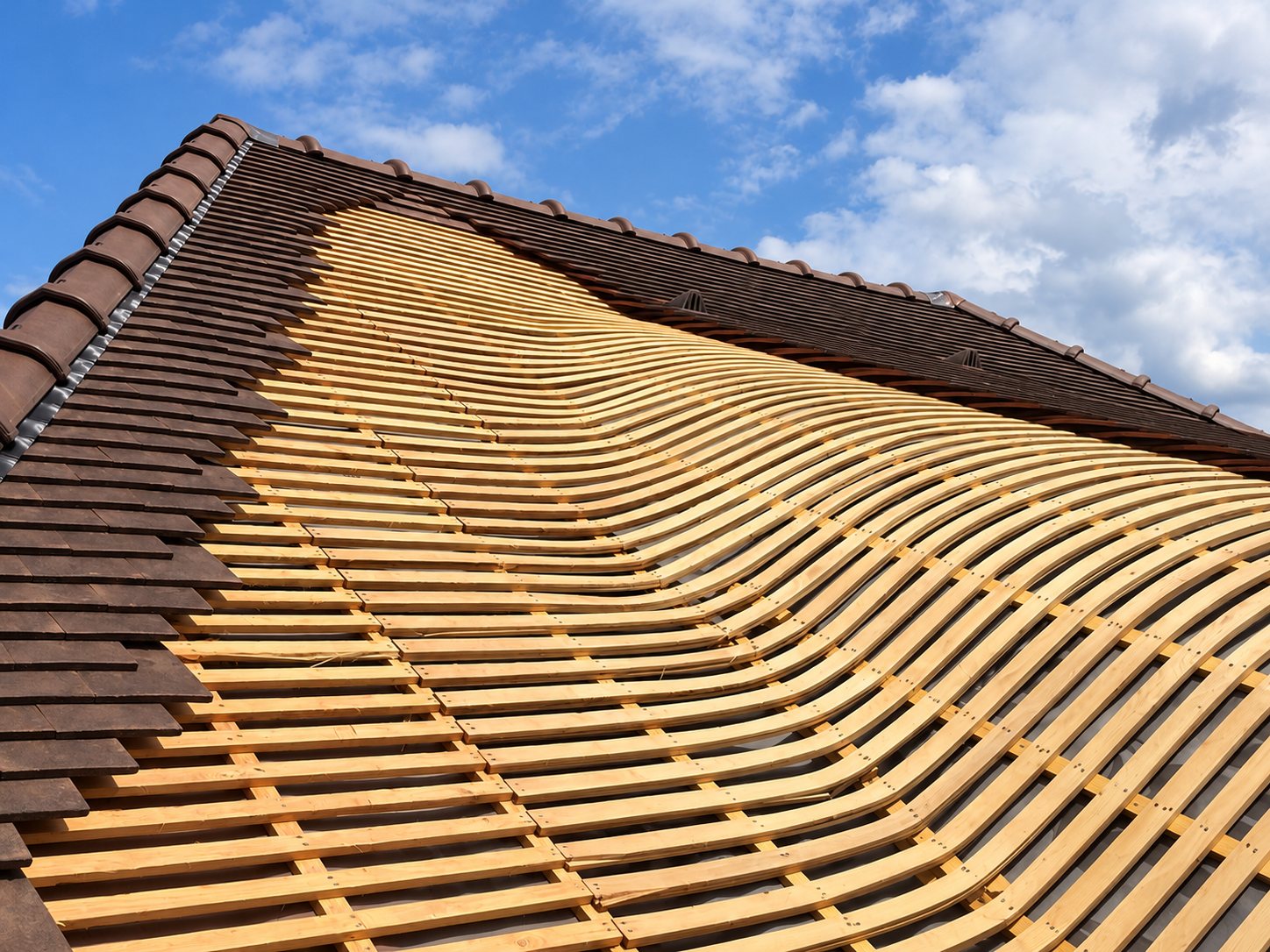 Vue en contre-plongée d'un toit dont les liteaux de bois dessinent une forme ondulée, bordé de tuiles brunes déjà posées sous un ciel bleu nuageux.