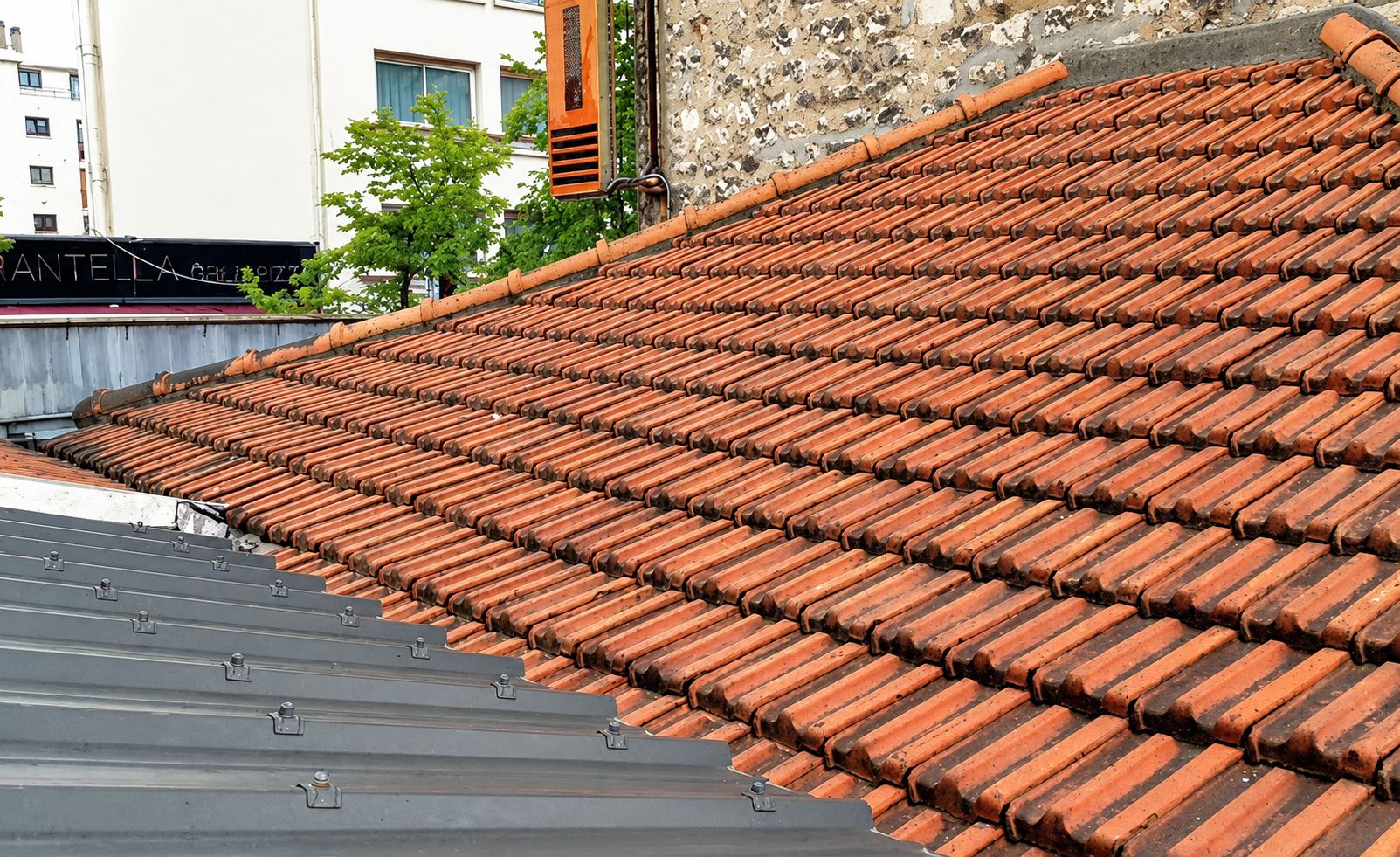 Toit en tuiles mécaniques rouges neuves bordé d'une couverture en bac acier gris, avec des immeubles et des arbres à l'arrière-plan.