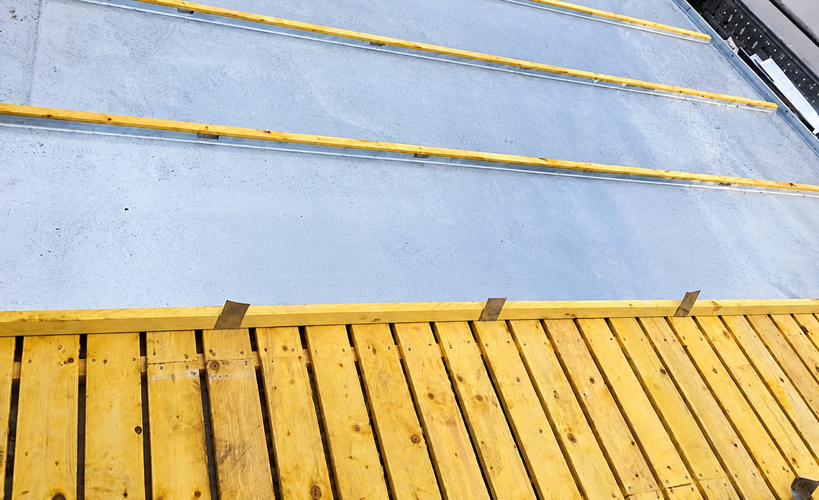 Vue rapprochée d'un toit en chantier avec des bandes de zinc grises séparées par des tasseaux en bois et un platelage en planches au premier plan.
